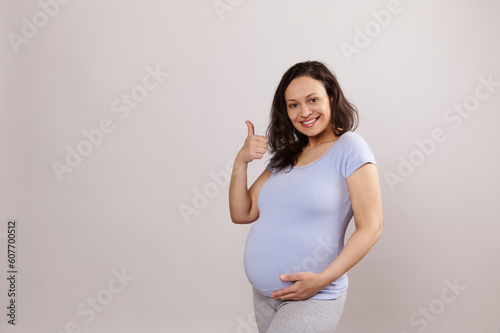 Multi ethnic beautiful pregnant woman, expectant mother in blue t-shirt, gently touching caressing her big belly and showing thumb up, smiling looking at camera, isolated background. Copy ad space