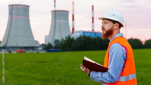 An engineer with a notebook on the background of a power plant. The man is wearing a white helmet and an orange vest