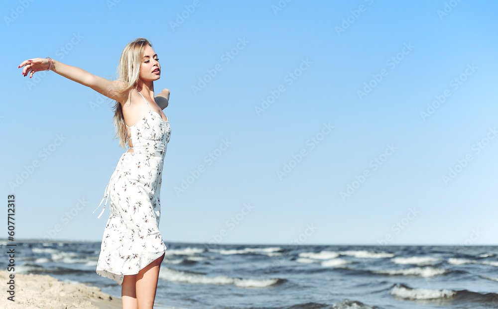 Happy blonde beautiful woman on the ocean beach standing in a white summer dress, open arms.