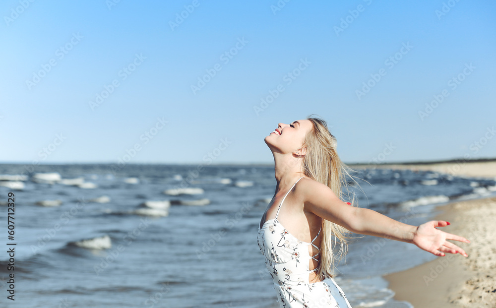 Happy blonde beautiful woman on the ocean beach standing in a white summer dress, raising hands.