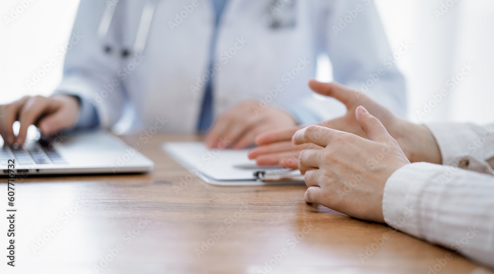 Doctor and patient sitting at the wooden desk in clinic. Female physician's hands pointing into laptop computer monitor, close up. Medicine concept. Doctor and patient sitting at the wooden desk in clinic. Female physician's hands pointing into laptop computer monitor, close up. Medicine concept.