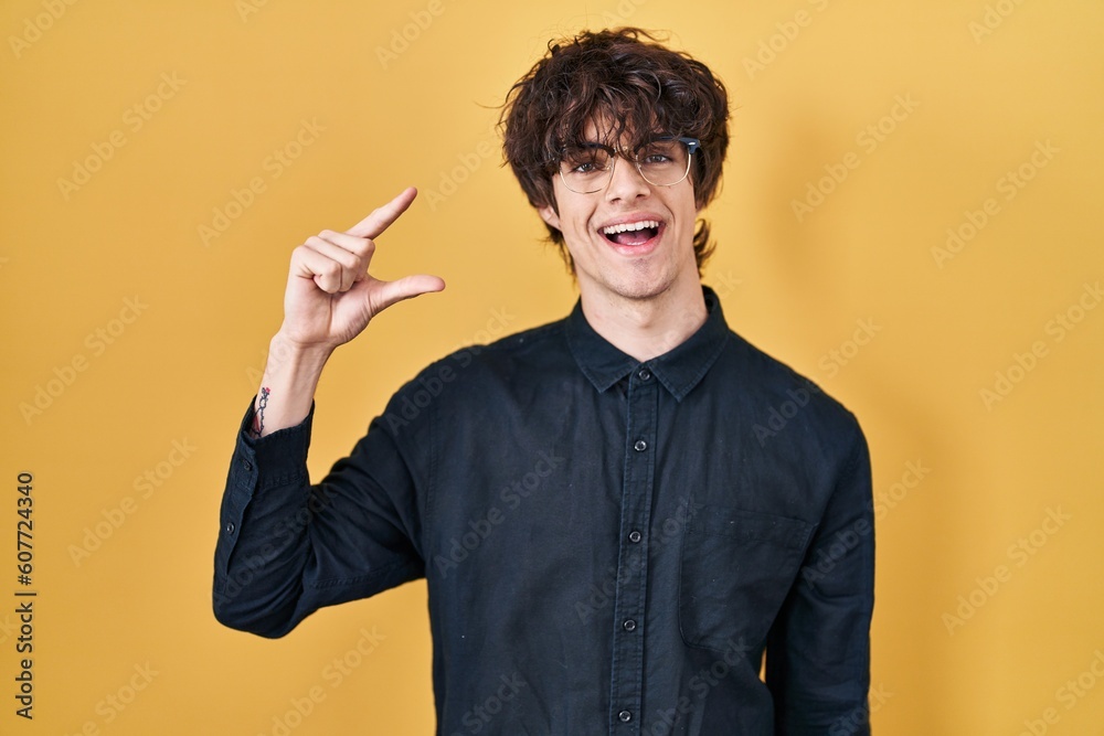 Young man wearing glasses over yellow background smiling and confident gesturing with hand doing small size sign with fingers looking and the camera. measure concept. Young man wearing glasses over yellow background smiling and confident gesturing with hand doing small size sign with fingers looking and the camera. measure concept.