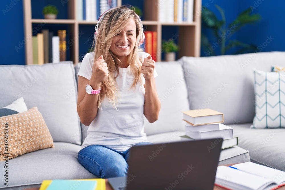 Young blonde woman studying using computer laptop at home excited for success with arms raised and eyes closed celebrating victory smiling. winner concept. Young blonde woman studying using computer laptop at home excited for success with arms raised and eyes closed celebrating victory smiling. winner concept.