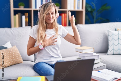Young blonde woman studying using computer laptop at home smiling swearing with hand on chest and fingers up, making a loyalty promise oath