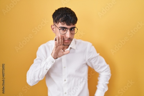 Young hispanic man standing over yellow background hand on mouth telling secret rumor, whispering malicious talk conversation