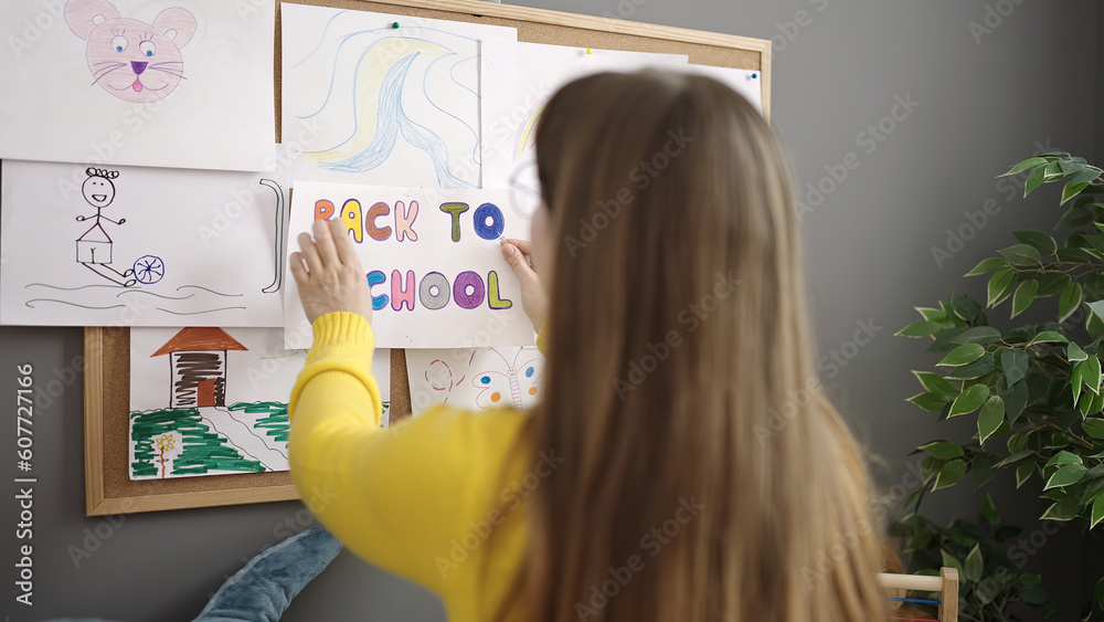 Young blonde woman preschool teacher hanging back to school paper on corkboard at kindergarten Young blonde woman preschool teacher hanging back to school paper on corkboard at kindergarten