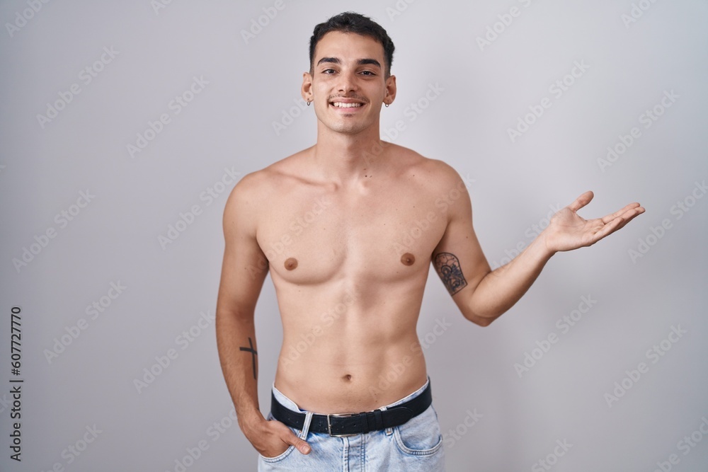 Handsome hispanic man standing shirtless smiling cheerful presenting and pointing with palm of hand looking at the camera.