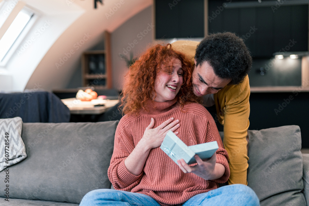 Red-haired woman with curly hair sitting on the couch and using her phone, her boyfriend came up behind he has a present for her