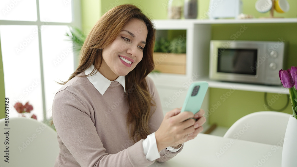 Young beautiful hispanic woman using smartphone sitting on table at home Young beautiful hispanic woman using smartphone sitting on table at home