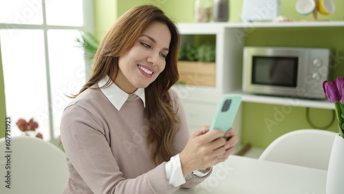 Young beautiful hispanic woman using smartphone sitting on table at home