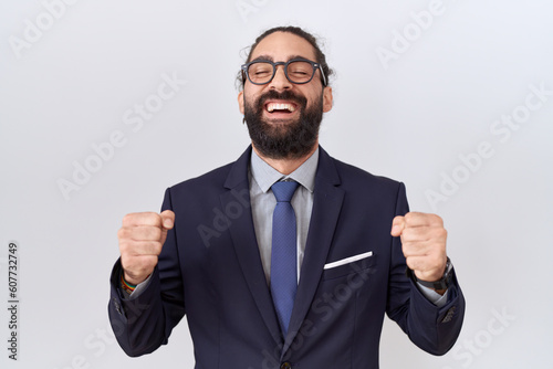 Hispanic man with beard wearing suit and tie excited for success with arms raised and eyes closed celebrating victory smiling. winner concept.