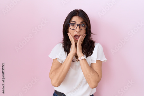 Middle age hispanic woman wearing casual white t shirt and glasses afraid and shocked, surprise and amazed expression with hands on face