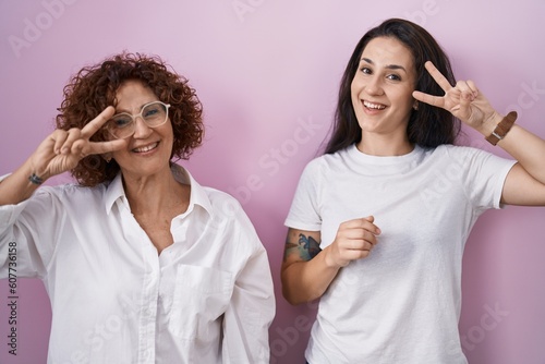 Hispanic mother and daughter wearing casual white t shirt over pink background doing peace symbol with fingers over face, smiling cheerful showing victory