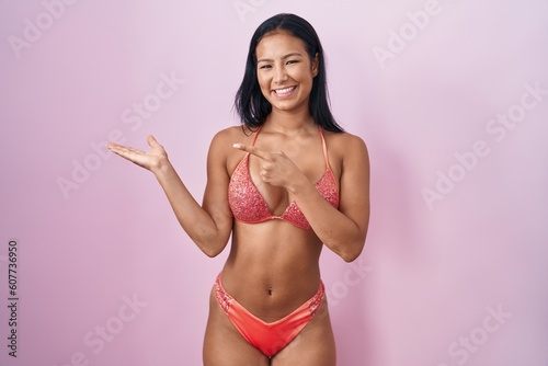 Hispanic woman wearing bikini amazed and smiling to the camera while presenting with hand and pointing with finger.