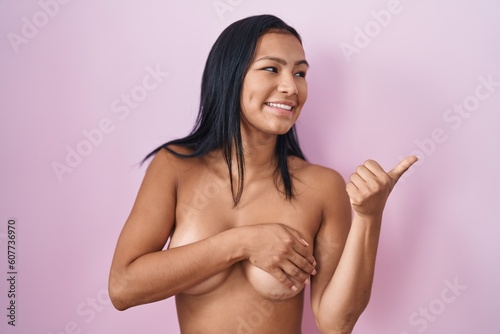 Hispanic woman standing topless covering breast with arm pointing thumb up to the side smiling happy with open mouth