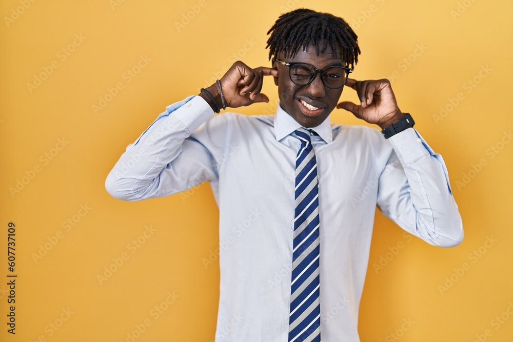 African man with dreadlocks standing over yellow background covering ears with fingers with annoyed expression for the noise of loud music. deaf concept. African man with dreadlocks standing over yellow background covering ears with fingers with annoyed expression for the noise of loud music. deaf concept.