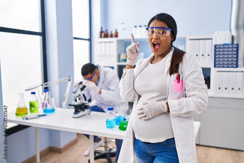 Young hispanic woman expecting a baby working at scientist laboratory pointing finger up with successful idea. exited and happy. number one. Young hispanic woman expecting a baby working at scientist laboratory pointing finger up with successful idea. exited and happy. number one.