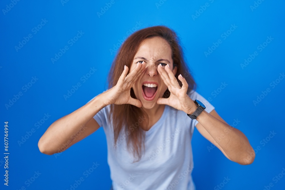 Fototapeta premium Brunette woman standing over blue background shouting angry out loud with hands over mouth
