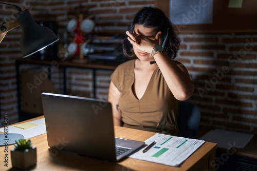 Young hispanic woman working at the office at night covering eyes with hand, looking serious and sad. sightless, hiding and rejection concept