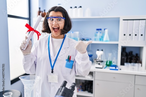 Young hispanic woman working at scientist laboratory holding degree pointing thumb up to the side smiling happy with open mouth