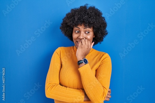 Black woman with curly hair standing over blue background looking stressed and nervous with hands on mouth biting nails. anxiety problem.