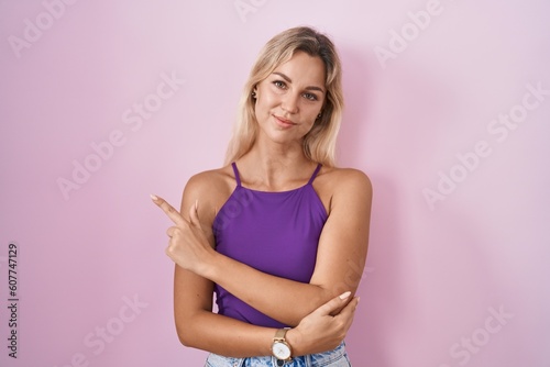 Young blonde woman standing over pink background pointing with hand finger to the side showing advertisement, serious and calm face © Krakenimages.com