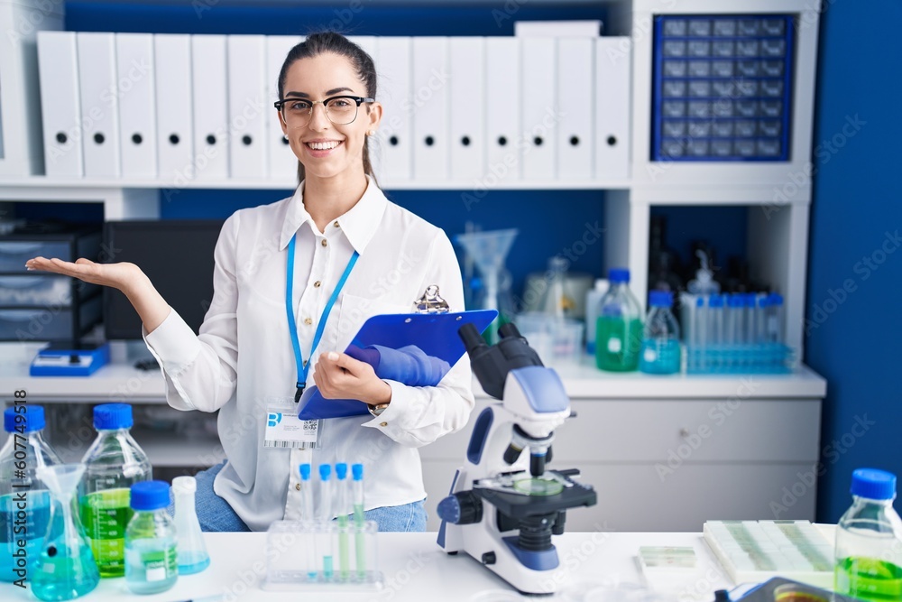 Young brunette woman working at scientist laboratory smiling cheerful presenting and pointing with palm of hand looking at the camera.