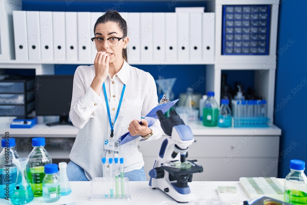 Young brunette woman working at scientist laboratory looking stressed and nervous with hands on mouth biting nails. anxiety problem.