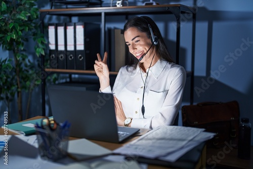 Young brunette woman wearing call center agent headset working late at night smiling with happy face winking at the camera doing victory sign. number two.