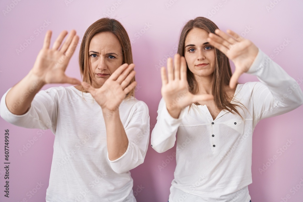 Middle age mother and young daughter standing over pink background doing frame using hands palms and fingers, camera perspective Middle age mother and young daughter standing over pink background doing frame using hands palms and fingers, camera perspective