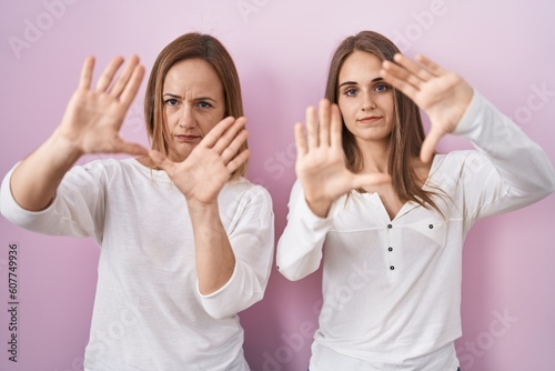 Middle age mother and young daughter standing over pink background doing frame using hands palms and fingers, camera perspective