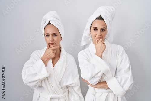Middle age woman and daughter wearing white bathrobe and towel looking confident at the camera with smile with crossed arms and hand raised on chin. thinking positive.