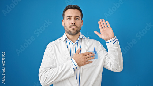 Young hispanic man doctor making an oath with hand on chest over isolated blue background © Krakenimages.com