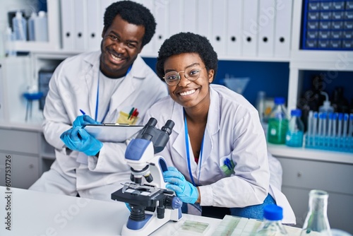 African american man and woman scientists using microscope writing on document at laboratory