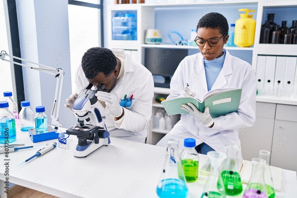 Man and woman scientists using microscope writing on notebook at laboratory Man and woman scientists using microscope writing on notebook at laboratory