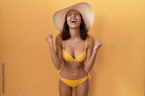 Young hispanic woman wearing bikini and summer hat celebrating surprised and amazed for success with arms raised and eyes closed. winner concept.