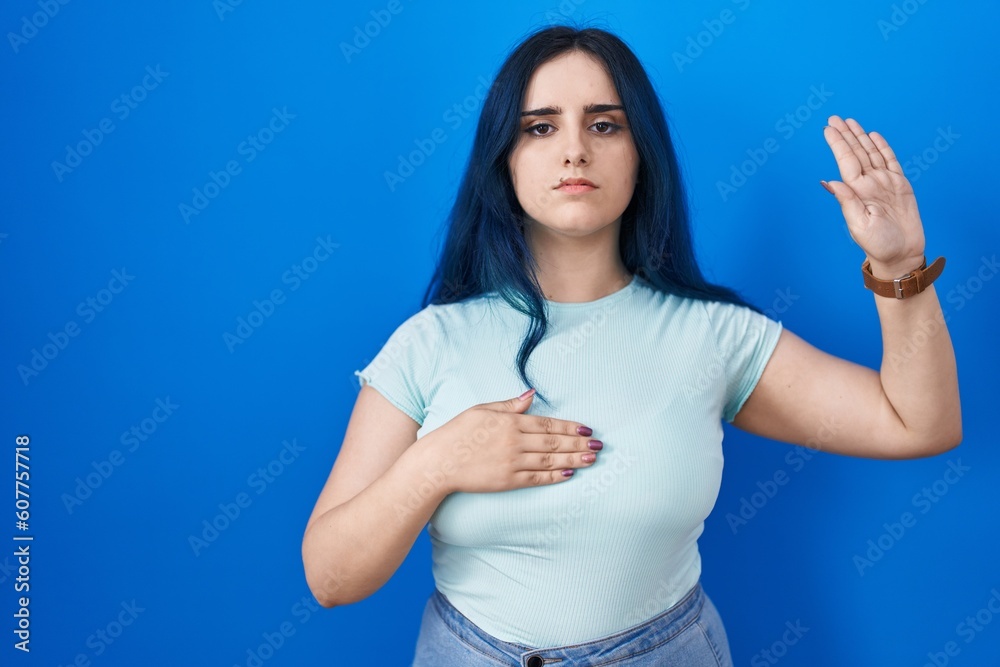 Young modern girl with blue hair standing over blue background swearing with hand on chest and open palm, making a loyalty promise oath Young modern girl with blue hair standing over blue background swearing with hand on chest and open palm, making a loyalty promise oath