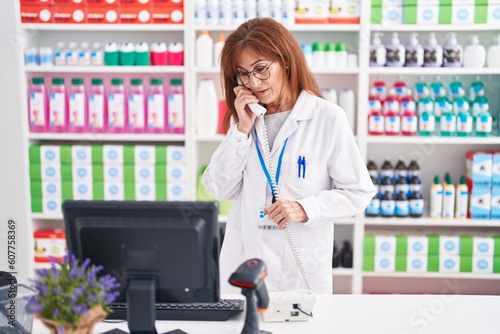 Middle age woman pharmacist talking on telephone using computer at pharmacy
