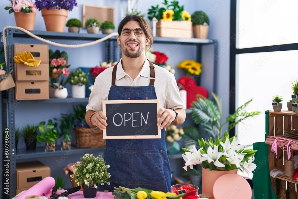 Hispanic man with long hair working at florist holding open sign sticking tongue out happy with funny expression. Hispanic man with long hair working at florist holding open sign sticking tongue out happy with funny expression.