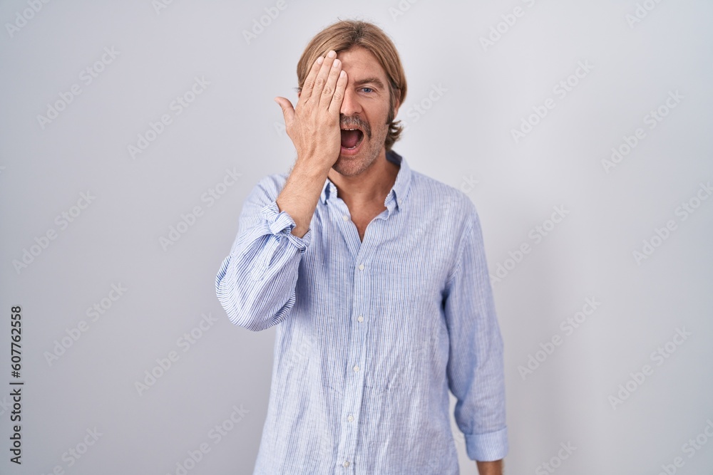 Caucasian man with mustache standing over white background covering one eye with hand, confident smile on face and surprise emotion.