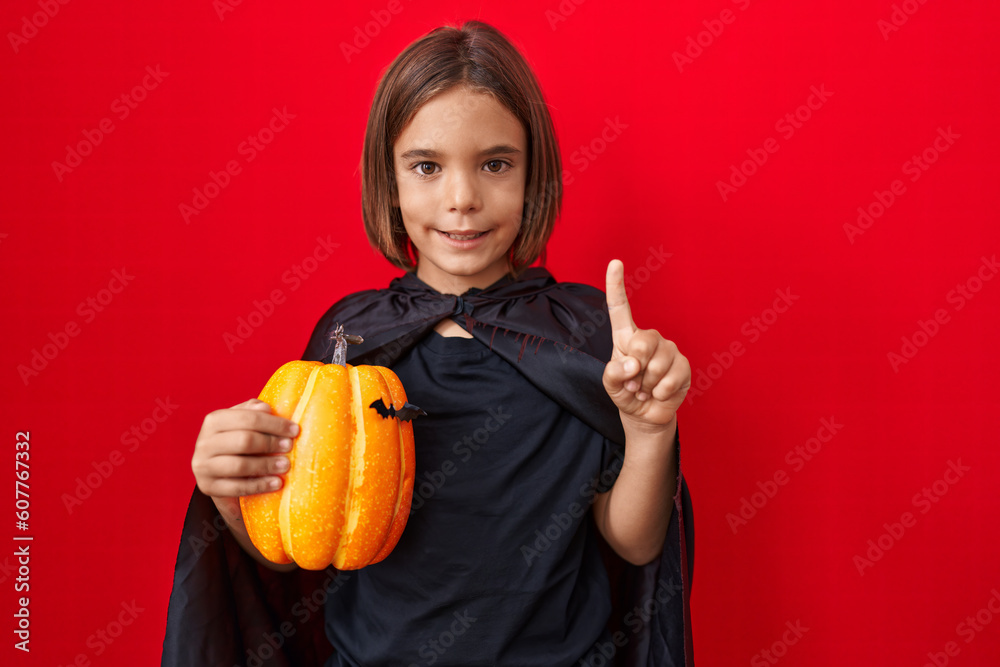 Little hispanic boy wearing a cape and holding halloween pumpkin pointing thumb up to the side smiling happy with open mouth Little hispanic boy wearing a cape and holding halloween pumpkin pointing thumb up to the side smiling happy with open mouth