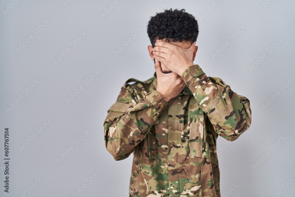 Arab man wearing camouflage army uniform covering eyes and mouth with hands, surprised and shocked. hiding emotion
