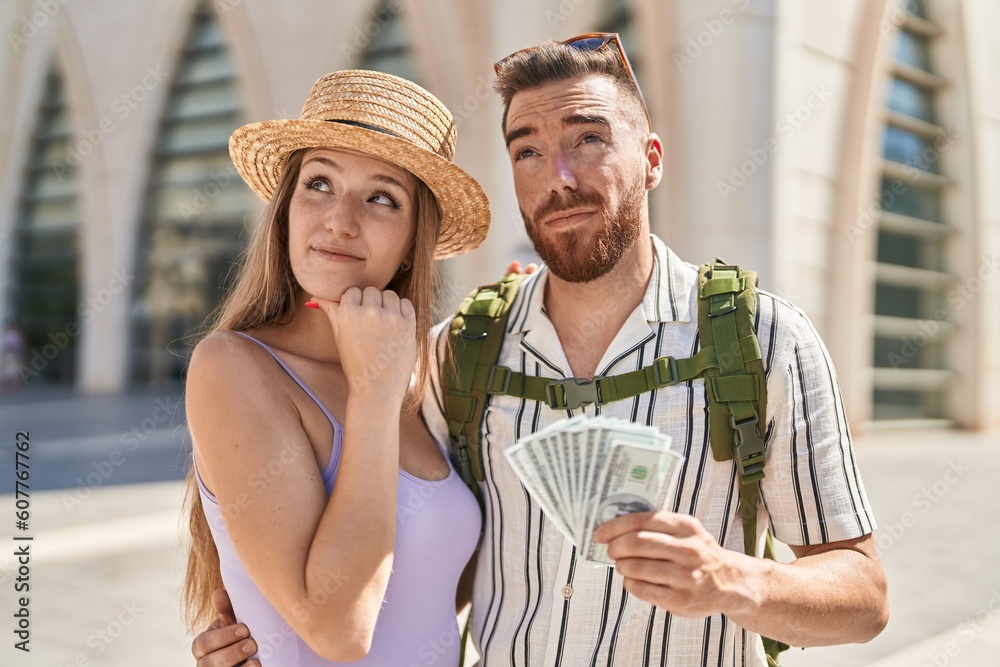 Young tourist couple holding dollars banknotes serious face thinking about question with hand on chin, thoughtful about confusing idea