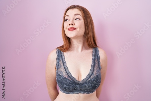 Redhead woman wearing lingerie over pink background smiling looking to the side and staring away thinking.