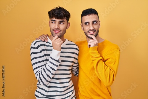 Young hispanic gay couple standing over yellow background with hand on chin thinking about question  pensive expression. smiling and thoughtful face. doubt concept.
