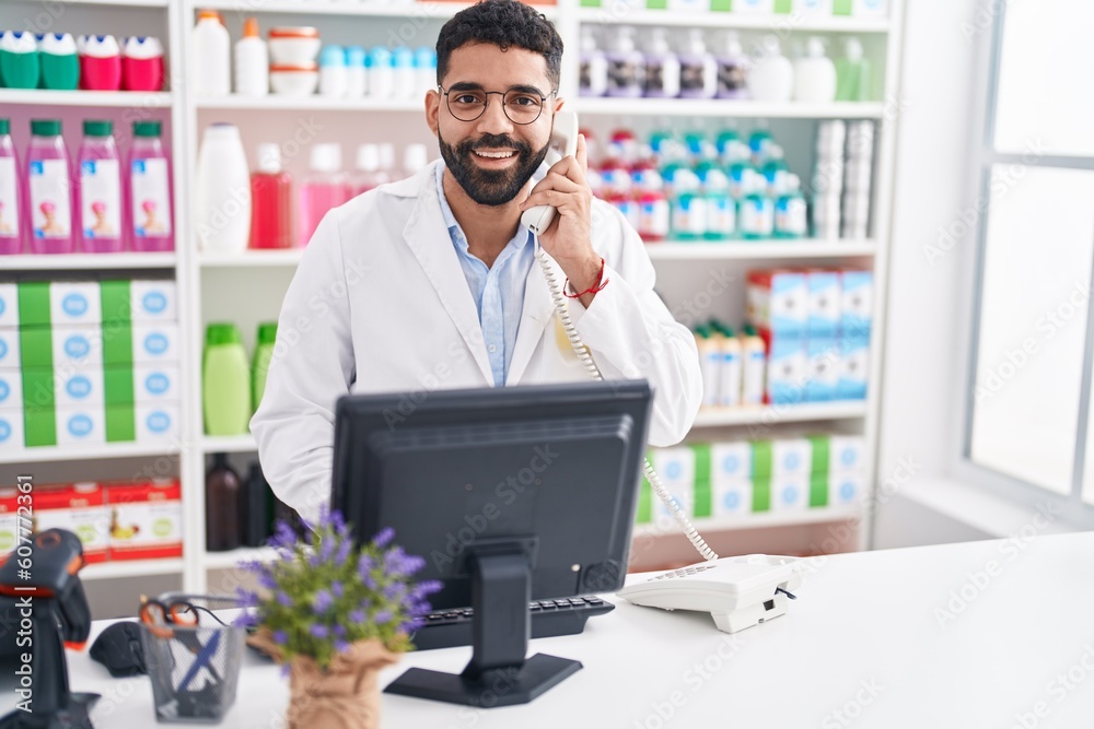Young arab man pharmacist talking on telephone using computer at pharmacy