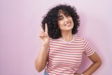 Young middle east woman standing over pink background smiling looking to the camera showing fingers doing victory sign. number two.