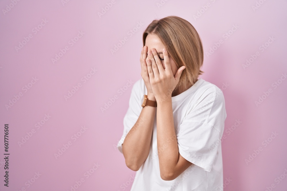 Young caucasian woman standing over pink background with sad expression covering face with hands while crying. depression concept.