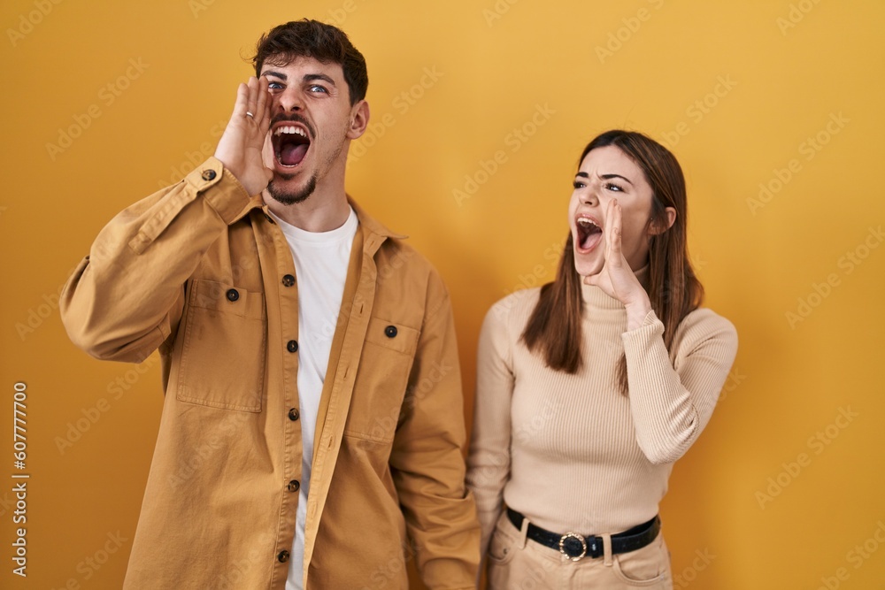 Young hispanic couple standing over yellow background shouting and screaming loud to side with hand on mouth. communication concept. Young hispanic couple standing over yellow background shouting and screaming loud to side with hand on mouth. communication concept.