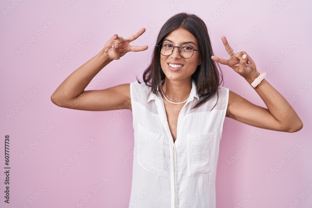 Brunette young woman standing over pink background wearing glasses doing peace symbol with fingers over face, smiling cheerful showing victory Brunette young woman standing over pink background wearing glasses doing peace symbol with fingers over face, smiling cheerful showing victory